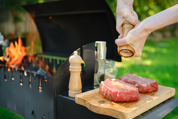 Middle-aged man sprinkles raw steak with pepper at backyard