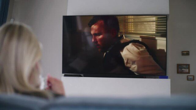 Adult woman sits on couch in cozy modern living room with cup of coffee, watches TV. Caucasian woman rests, spends weekend at home. Big flat screen TV monitor with displayed action movie on the wall.