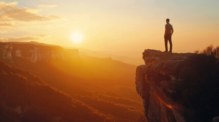 A young man stands on a cliff in the mountains as the sun sets, enjoying the beautiful view of nature.