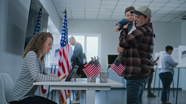 US citizen, male voter with kid comes to female polling officer for registration at polling station, takes paper bulletin for voting. National Election Day in the United States of America. Patriotism.