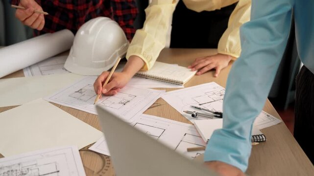 Architect and safety engineer discussion about building blueprint displaying on laptop on table with architectural document and wooden block scatter around at modern office. Closeup. Delineation.