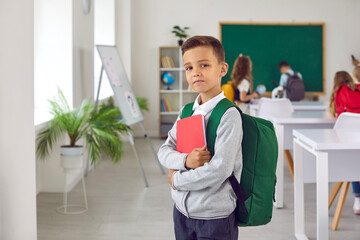 Portrait of a little school child. Schoolboy with a book and backpack. Clever elementary student boy with a bag and notebook standing in the classroom and looking at the camera. Back to school concept