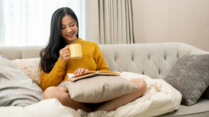 Fototapeta premium A beautiful, relaxed Asian woman is having a hot drink while reading a book on a sofa at home.