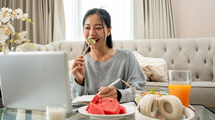 A woman is eating breakfast while working or studying online on her laptop at a coffee table.