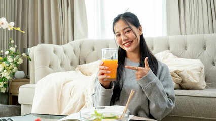 An attractive, smiling Asian woman is pointing her finger at a glass of orange juice in her hand.