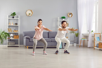 Obraz premium Happy sporty young couple wearing sportswear having sport workout in living room at home. Cheerful man and woman doing squats exercises together. Concept of family sport and fitness.