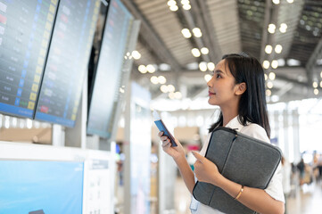 A beautiful Asian woman with her passport is standing in front of a flight information display.