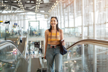 A gorgeous young Asian female tourist passenger is on an airport escalator, carrying her luggage.