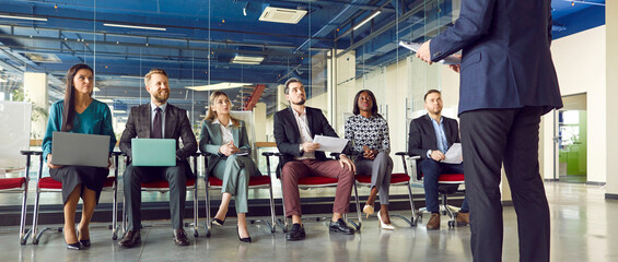 Man speaker talking on a work meeting in modern office in front of a small group of happy smiling company employees sitting on chairs in a row with laptops on a business conference. Banner.