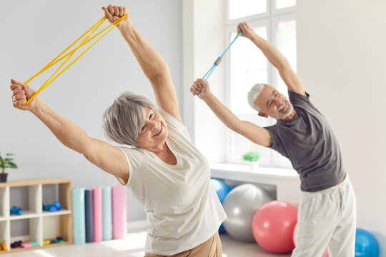 Smiling mature senior man and woman doing stretching exercises with rubber band standing in gym. Happy elderly people doing workout in rehabilitation center for health. Sport and fitness concept.