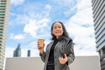 A confident Asian senior businesswoman stands in the urban city surrounded by skyscraper buildings.