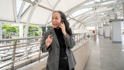 Obraz premium A confident mature Asian businesswoman is talking on the phone while standing on a skywalk.