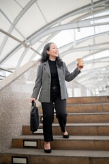 A happy mature Asian businesswoman is walking down the stairs on a skywalk in the urban city.