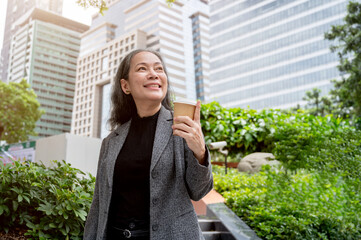 Fototapeta premium Positive mature Asian businesswoman is looking up at the skyscraper while standing in the city park.