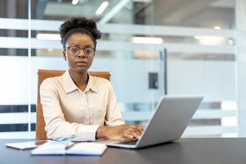 Confident businesswoman with glasses sitting at desk, working on laptop in modern office. Professional attire, focused expression, depicts dedication and productivity in corporate environment