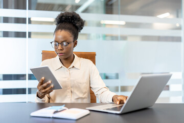 Professional businesswoman using tablet and laptop in modern office. Focused on work, demonstrating multitasking and productivity. Office furniture and glass partitions visible in background.