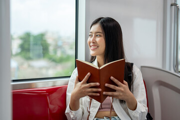 A happy Asian woman is looking at the view outside and reading a book while taking a sky train.