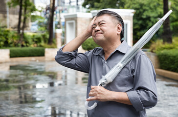 Ill Asian middle-aged man holding umbrella after raining, sick Asian uncle having cold, flu, fever,...