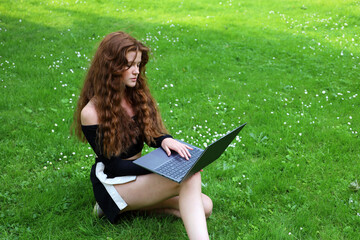 Attractive girl with long ginger hair sitting with laptop on a grass in summer park working outdoor