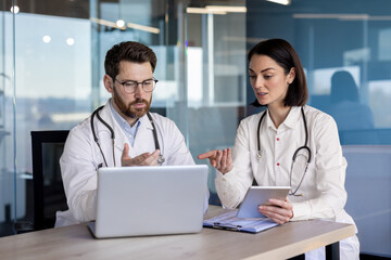Medical professionals in discussion about patient information using laptop and tablet in modern office setting. Team of healthcare workers collaborating, consulting, and reviewing medical data.