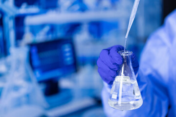 A person is holding a beaker filled with a clear liquid. The person is wearing a lab coat and gloves. Concept of scientific experimentation and precision