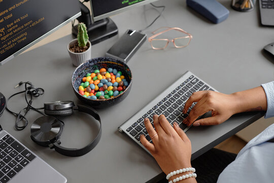 High-angle shot of gray working desk with candy bowl, headphones and hard drive placed on table while hands of female computer programmer typing source codes on keyboard in office