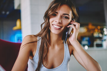 Fototapeta premium Portrait of cheerful hipster girl smiling at camera while making notes and calling on smartphone device.Positive young woman laughing during mobile conversation on cellular in cafe interior