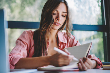 Obraz premium Pensive young businesswoman making notes of working plan in notepad sitting at laptop computer in coworking space.Hipster student writing homework and preparing for studying seminar at netbook