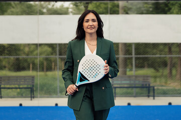 Confident businesswoman in green suit holding a padel tennis racket on a blue court. Outdoor...