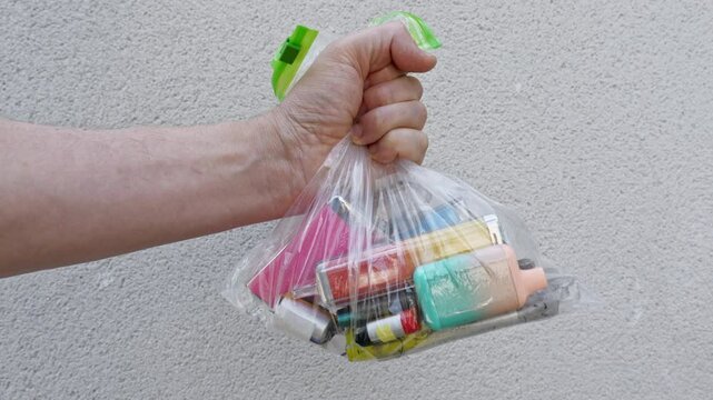 A white male grips a clear plastic bag with a collection of discarded vapes inside. The bag is held in front of a pale coloured stone wall.