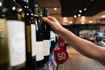 A person selects a bottle of wine from a supermarket shelf, with a focus on their hand and the wine bottles. male consumer concept 