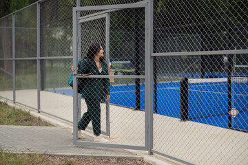 Woman in green suit entering a padel tennis court through a chain-link gate. The court has a vibrant blue surface.