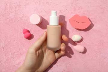 Bottle with foundation in hand and cosmetic sponges on pink background, top view