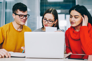 Successful hipster student rewriting information from business card in search job.Positive young people dressed in casual wear collaborating at laptop computer in office sitting at modern laptop