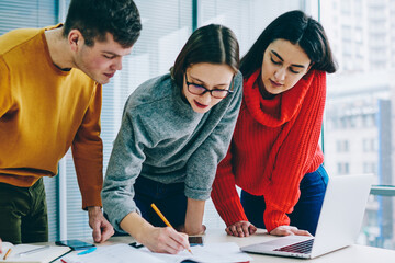 Pensive hipster students working in team and drawing sketch standing over desktop with laptop in...