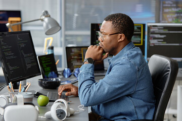 Side view of thoughtful male African American VR programmer writing code for cutting-edge game development at working desk with multiple computer screens in office, copy space