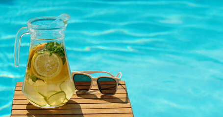 A glass pitcher with refreshing lemon and mint water on a wooden table beside white sunglasses, set...
