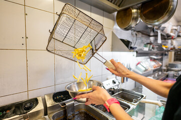 Cook draining freshly fried french fries from a metal basket in a commercial kitchen.