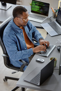 Vertical high-angle shot of concentrated male African American software developer writing program code fixing errors while sitting at working desk with laptops in office