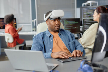 Portrait shot of software engineer of Black ethnicity in VR headset writing code for cutting-edge game development at working desk in office, copy space