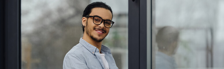 A cheerful man stands in front of a window.
