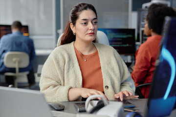 Portrait shot of concentrated higher-weight female Middle Eastern software developer working on computer writing program code while looking at screen sitting at desk in IT company office, copy space