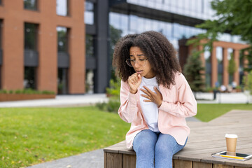Fototapeta premium Young woman coughing while sitting outside urban building. Appears unwell and holding chest, suggesting possible respiratory issue or cold. Outdoor park setting with trees and modern architecture