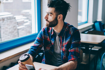 Thoughtful hipster guy looking at cafeteria window while waiting for call from friend on new smartphone and enjoying caffeine beverage, pensive man holding mobile phone and cup of coffee indoors