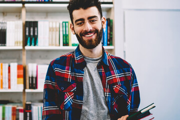 Half length portrait of smiling bearded male teenager selecting book in library of college standing near shelves, happy successful hipster guy picking literature and making research in bookstore