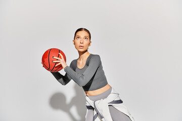 A sporty young woman in active wear holding a basketball in her right hand against a grey background.