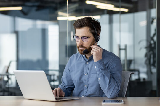 A young serious man works in the office on a laptop, wears a headset and talks on a video call, provides advice and support