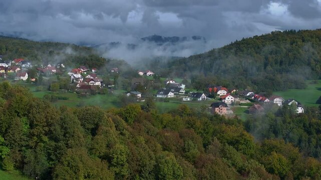 AERIAL: Charming village nestled among rolling hills, shrouded in morning mist. Lush forest in colorful shades of autumn surrounding an idyllic rural settlement in the hilly countryside on a sunny day