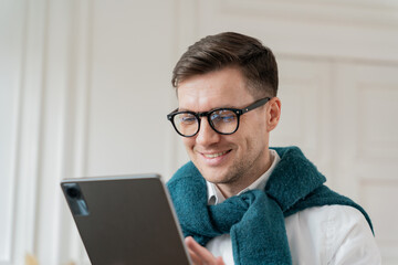 Smiling man in glasses with a sweater draped over shoulders, using a tablet in a bright, elegant...