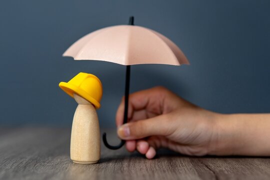 Closeup image of hand holding a umbrella over a wooden doll wearing a yellow hard hat. Worker and employee insurance concept.